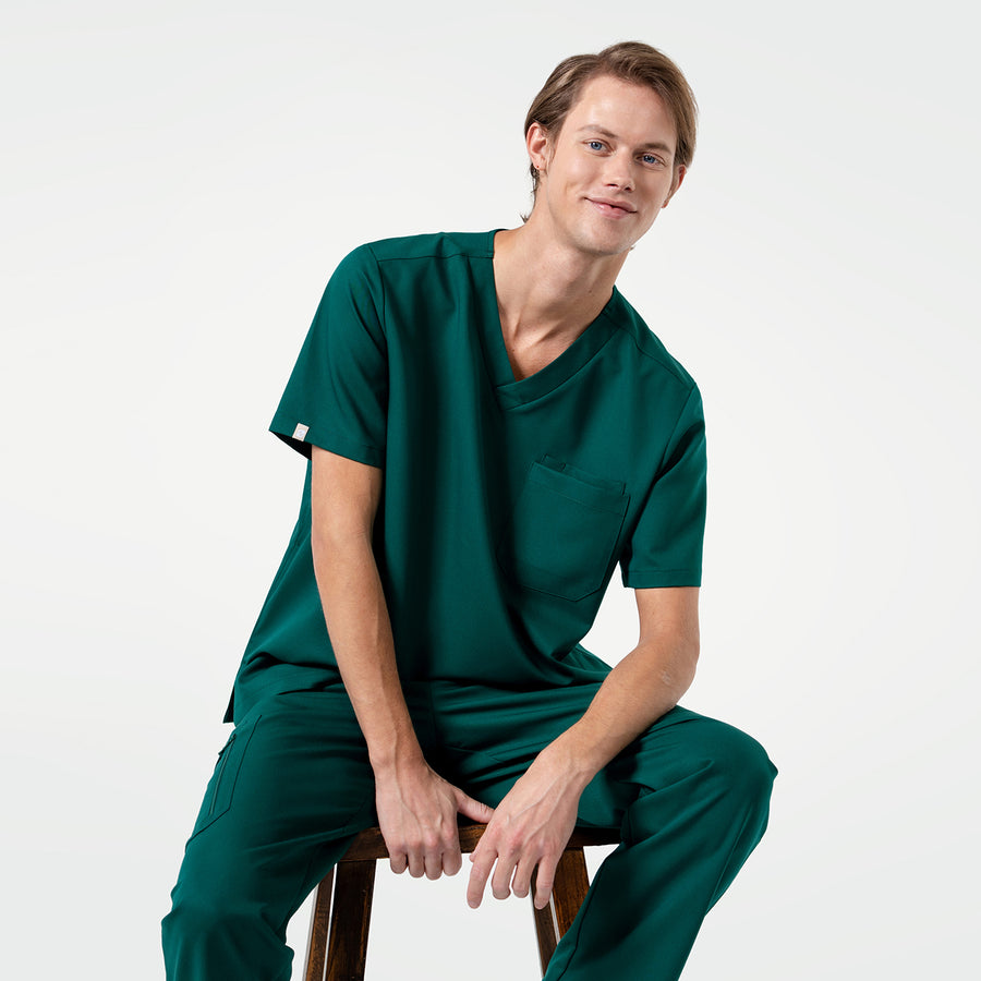 Wearing a Pine Green 3 Pocket Scrub Top, the model sits comfortably with a slight smile.