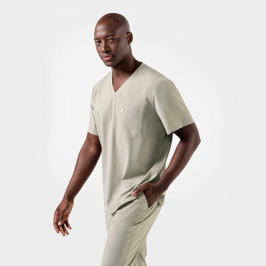Wearing the Sand Khaki Single Pocket Scrub Top, the model walks confidently with a composed look, hand in the pocket and body slightly angled.