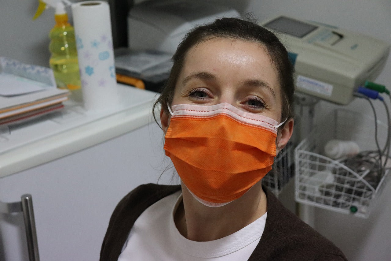 Image of Nurse in a surgery room with medical equipment behind her, smiling at the camera with a orange surgical mask on with a white shirt and black cardigan.