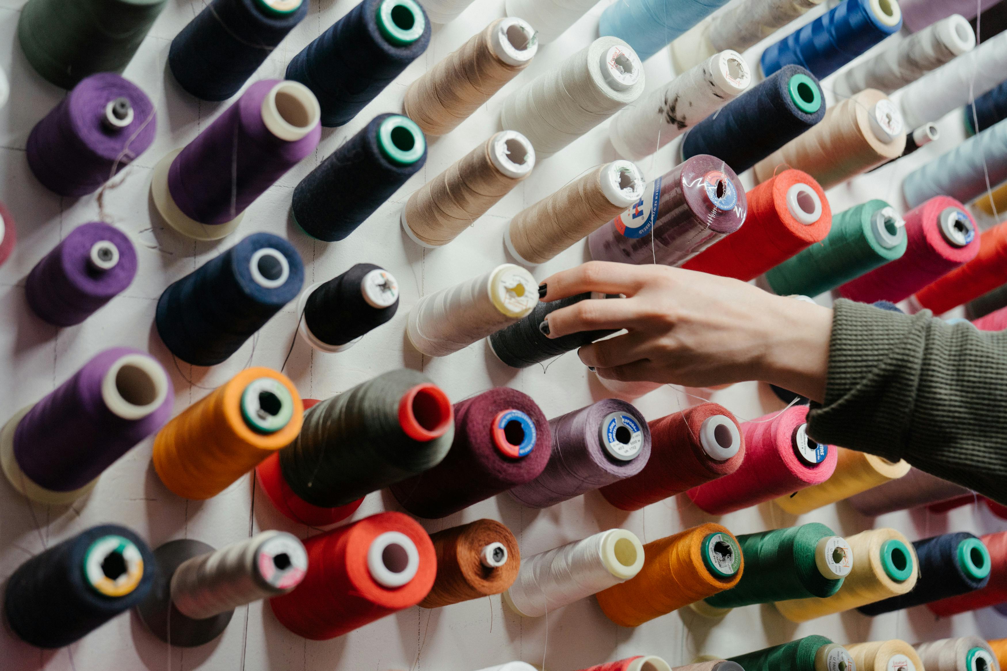 Image of A wall-mounted display of colorful sewing thread spools in various shades, with a hand reaching for one, showcasing a creative workspace.