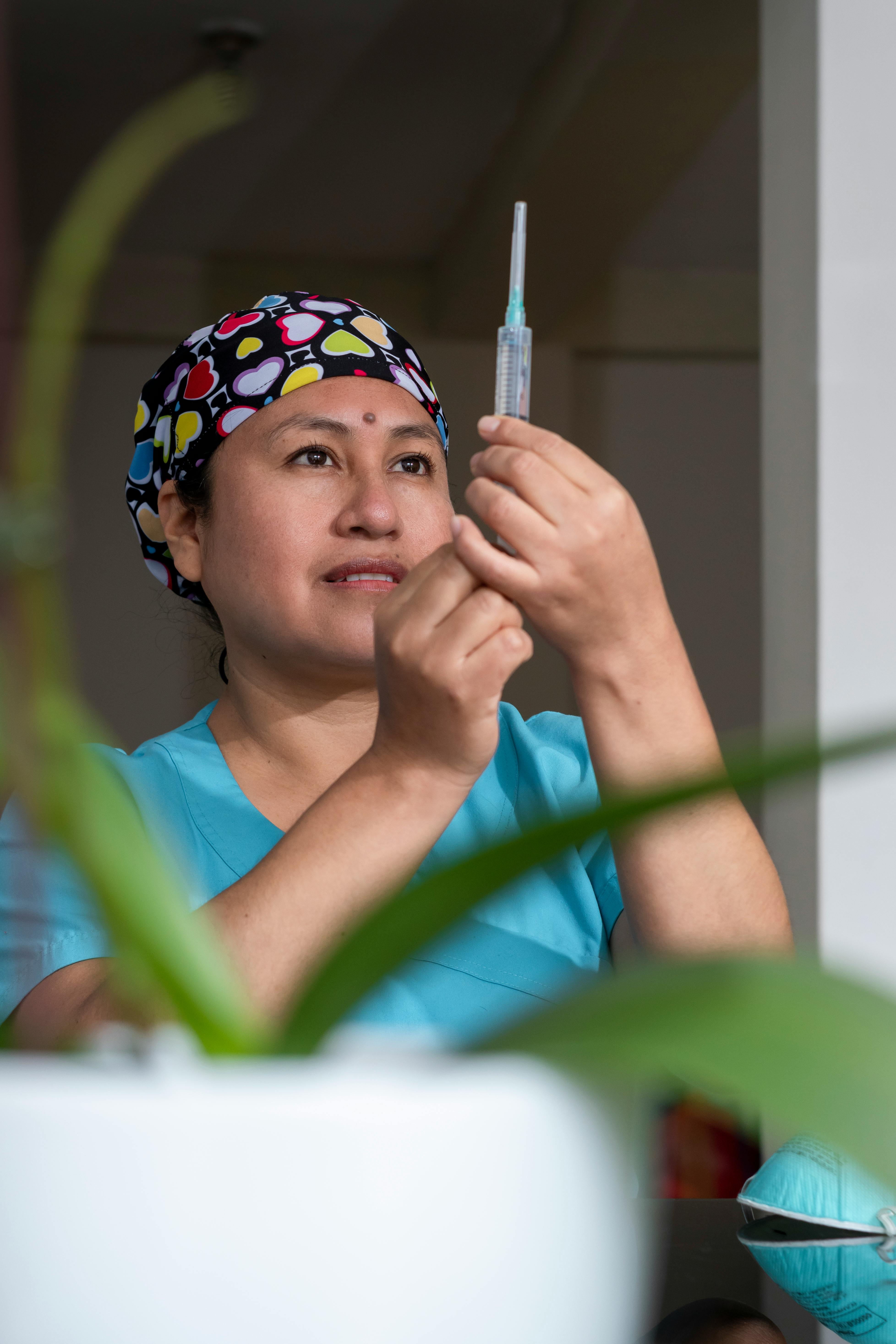 Image of A nurse in teal scrubs holding a syringe, wearing a colorful headband with heart patterns, preparing for a procedure.