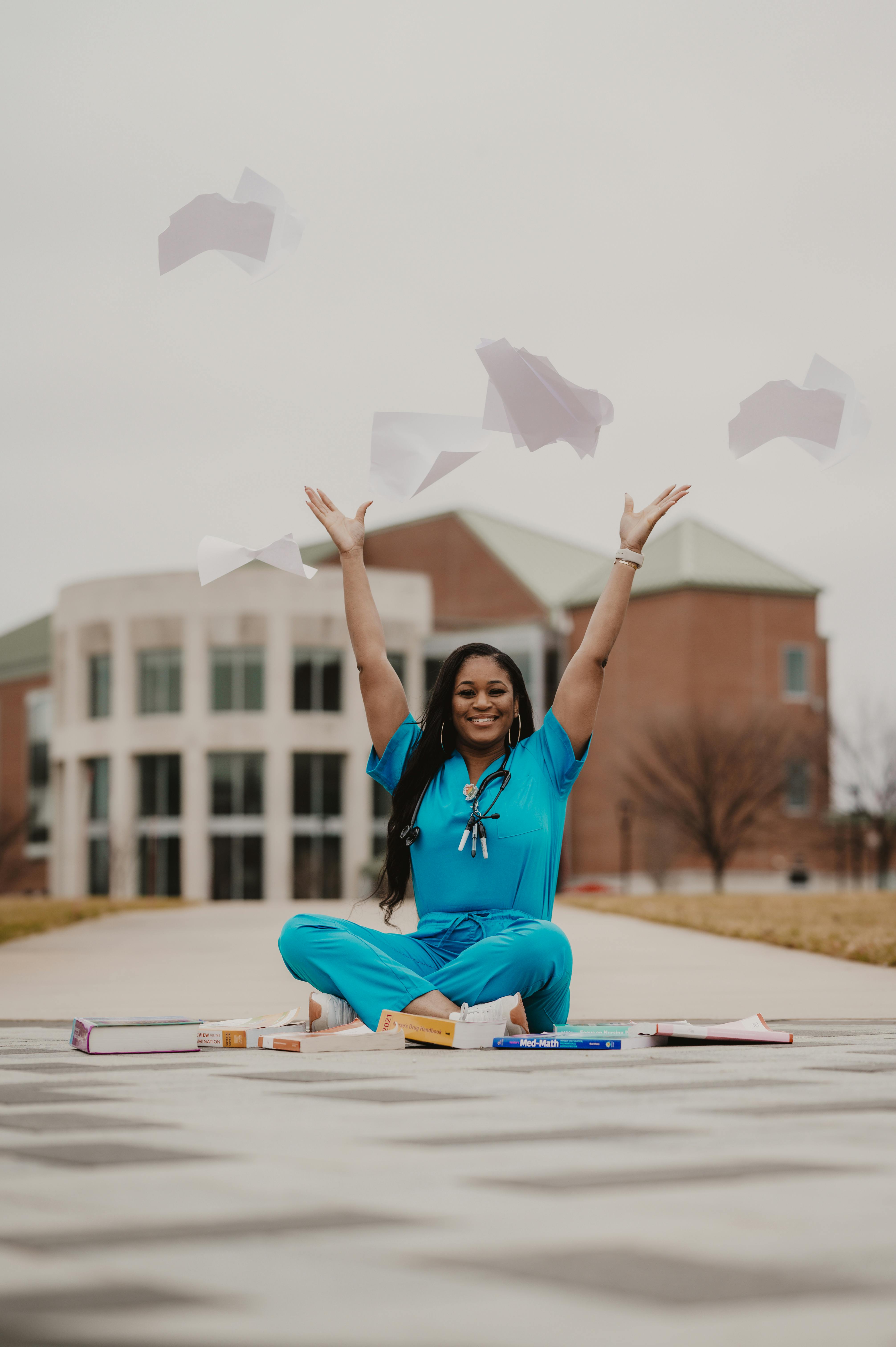 Image of Medical student sitting in front of campus building with blue scrub top and bottom on, throwing papers in the the air with books scattered around her.