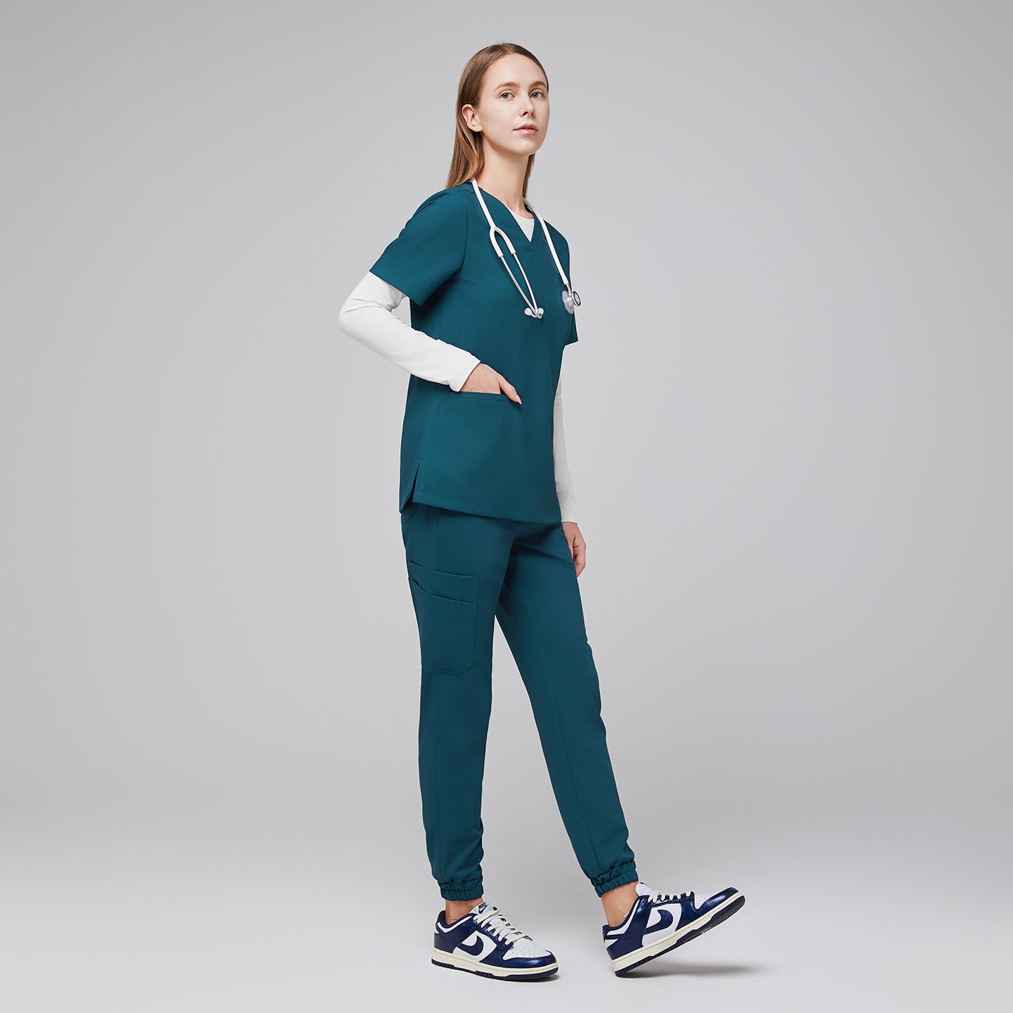 Image of Model facing forward wearing ocean teal scrub top and ocean teal jogger scrub pants, with a white underscrub and hand in pocket.