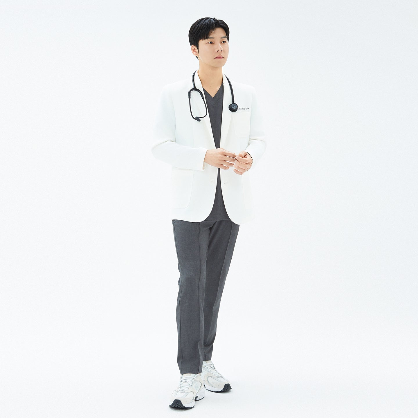 Man standing with white background with a white lab coat and a charcoal gray set of scrubs under.