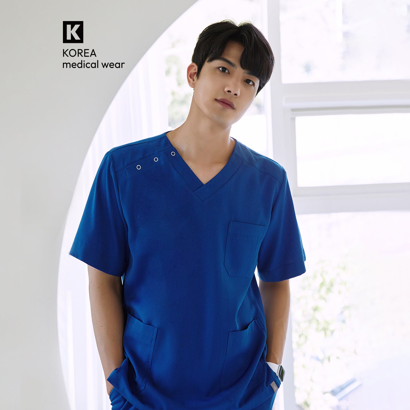 Image of Male model wearing a Royal Blue DS-167 scrub top, standing in a bright clinical setting with hands in pockets, showcasing the V-neck design and shoulder snap details.