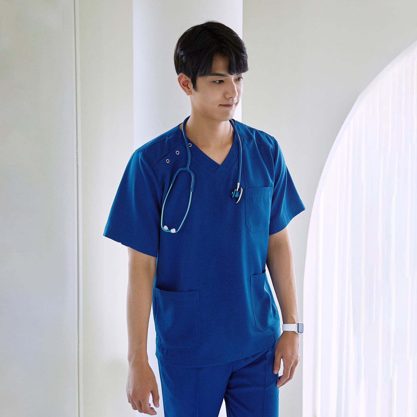 Image of Male model wearing a Royal Blue DS-167 scrub top with a stethoscope, standing in front of a bright window while highlighting the top’s chest pocket and functional lower pockets.