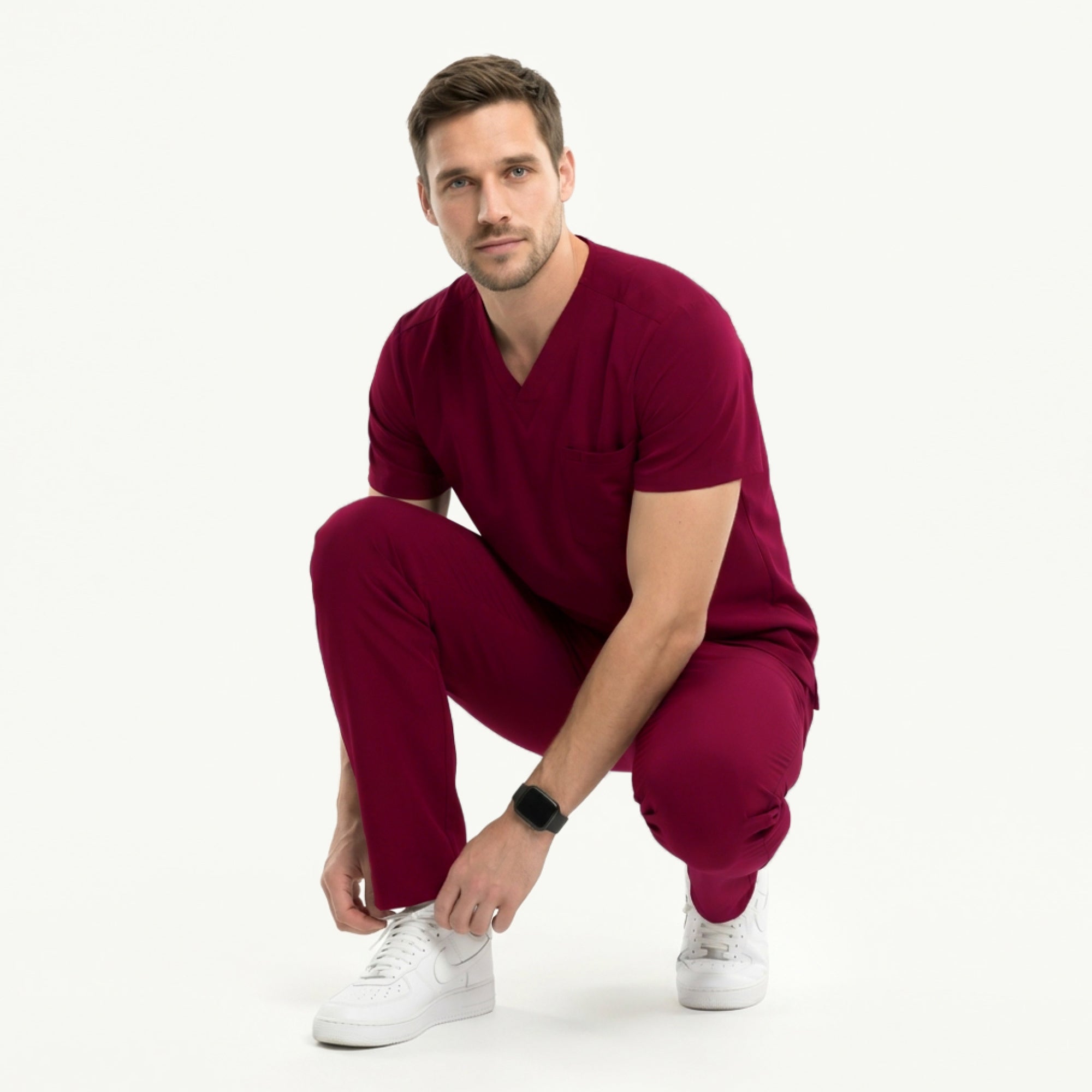 Image of Man wearing a burgundy V-neck scrub top with badge and pocket accessories, paired with matching burgundy scrub pants and white sneakers,Burgundy