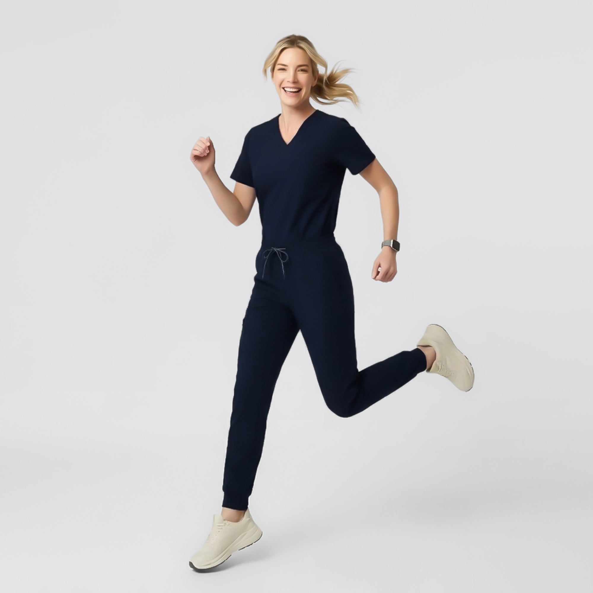 Image of Model walking in a navy V-neck pleated-shoulder scrub top and matching jogger scrub pants, hands in pockets, against a clean light background.