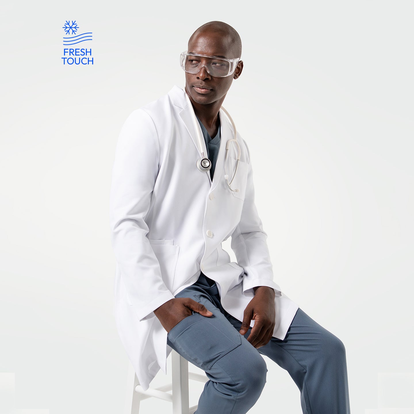 Image of Seated in the Men's LCM-101 Lab Coat in White over medical scrubs, with a stethoscope around his neck and protective glasses, looking to the side