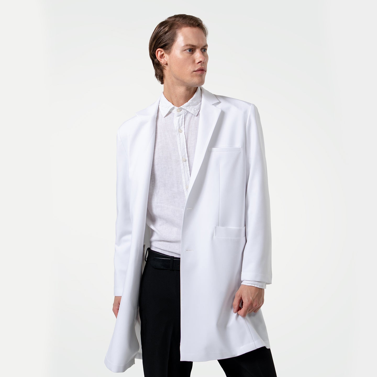 Image of Looking to the side, a male doctor wears a white lab coat over a white dress shirt and black pants on a clean light gray background.