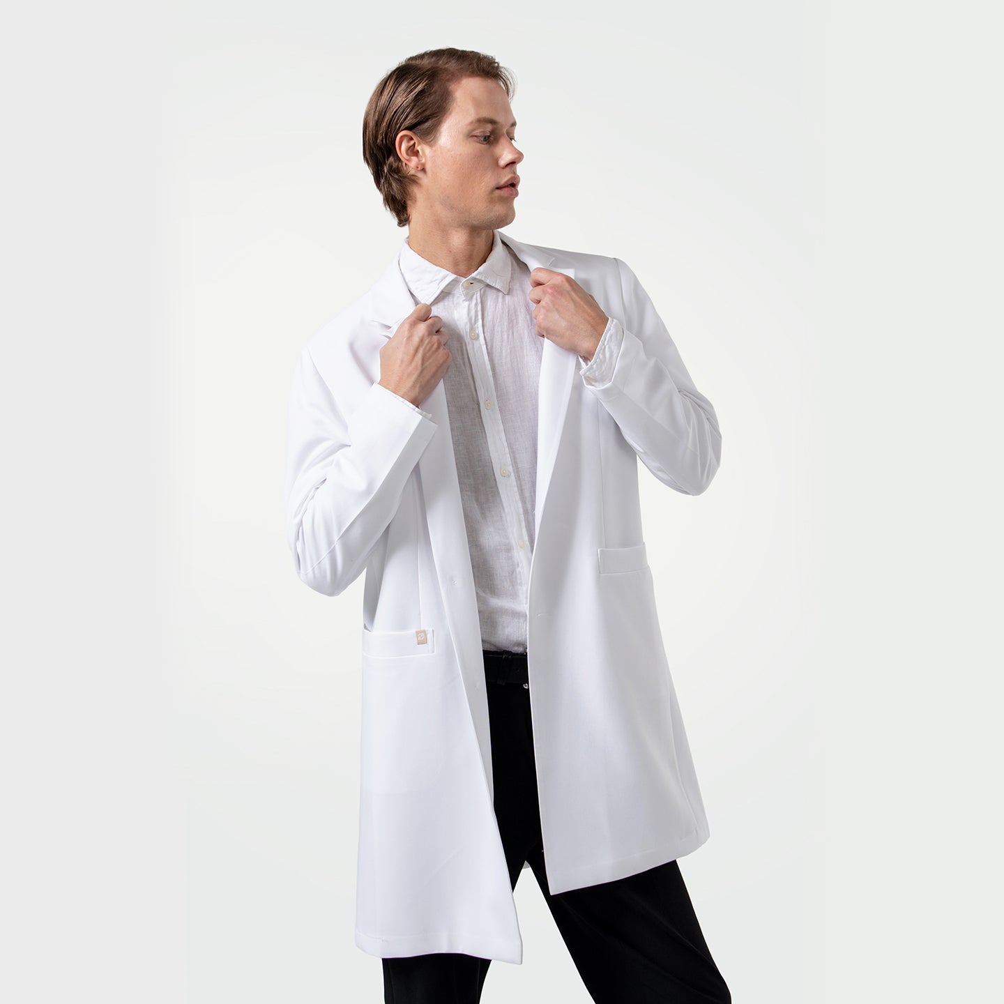 Image of Confident male healthcare professional wearing a white coat over a white dress shirt, adjusting the lapels of the coat while looking to the side on a clean white background.