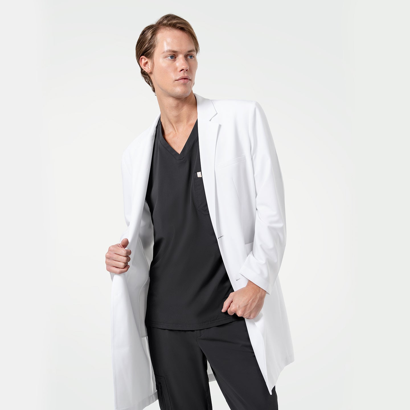 Image of Lcm-103 men’s white lab coat styled over black scrub top and pants, model holding coat open to show inner fit, professional medical apparel on light background.
