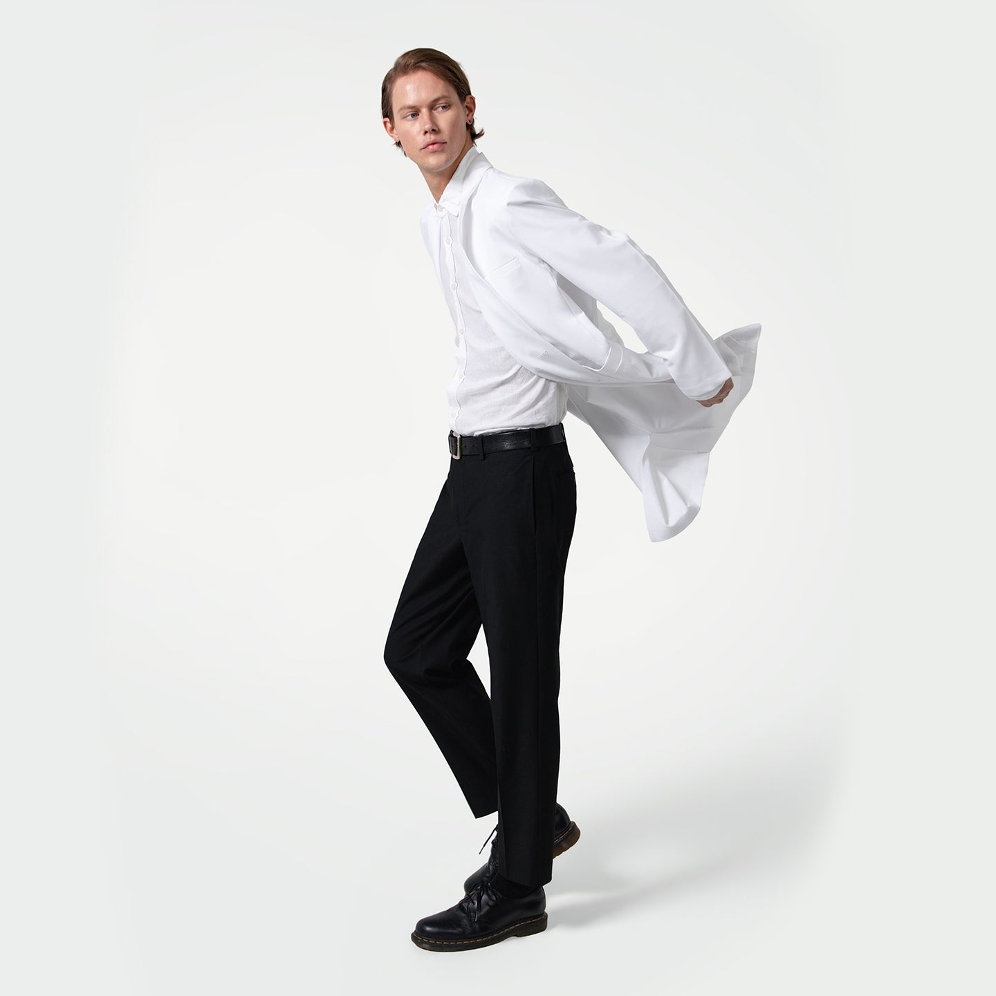 In a dynamic pose, a male healthcare professional slips into a white coat over a dress shirt and black pants on a clean light gray background.