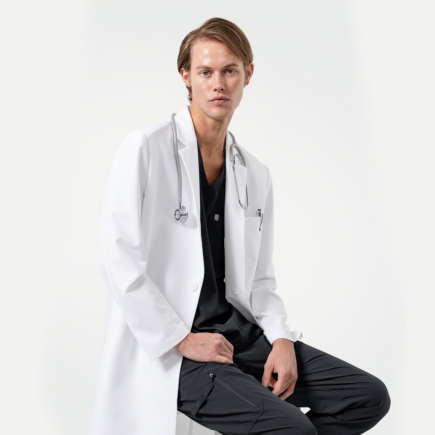 Image of Lcm-103 men’s white lab coat with stethoscope, styled over black scrubs, modern tailored design for doctors and healthcare professionals.