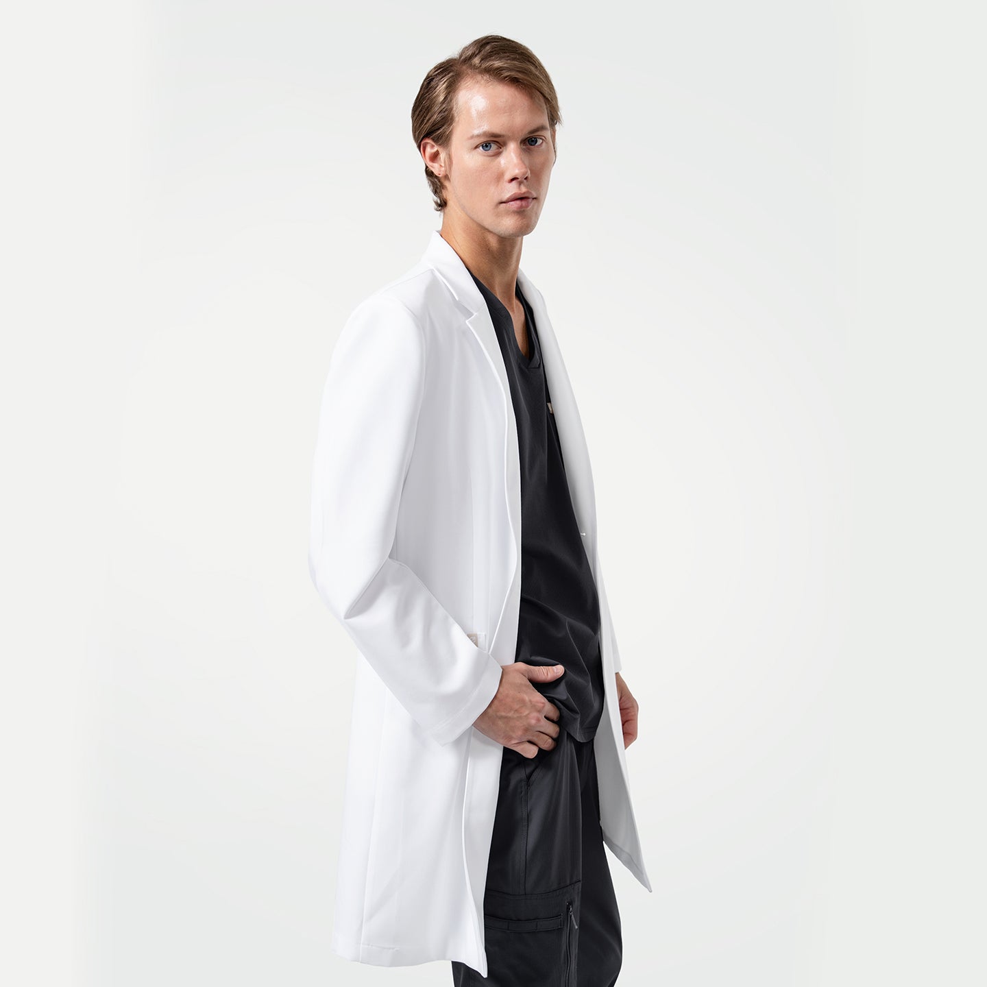 Image of Lcm-103 white lab coat for men, shown on model wearing black scrubs underneath, standing sideways with one hand in pocket against light background.