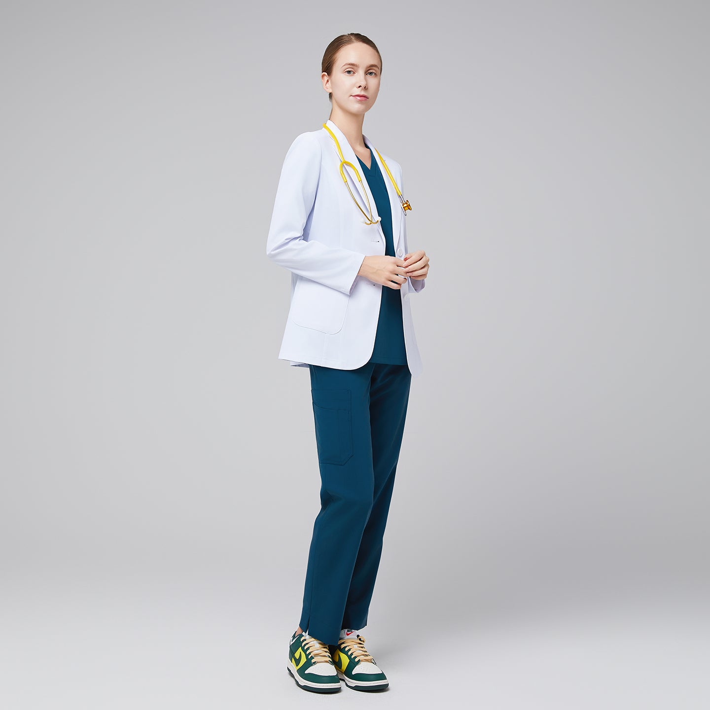 Model facing sideway wearing a white short lab coat, and blue scrub top and bottom under, with yellow stethoscope around her neck and green sneakers,White