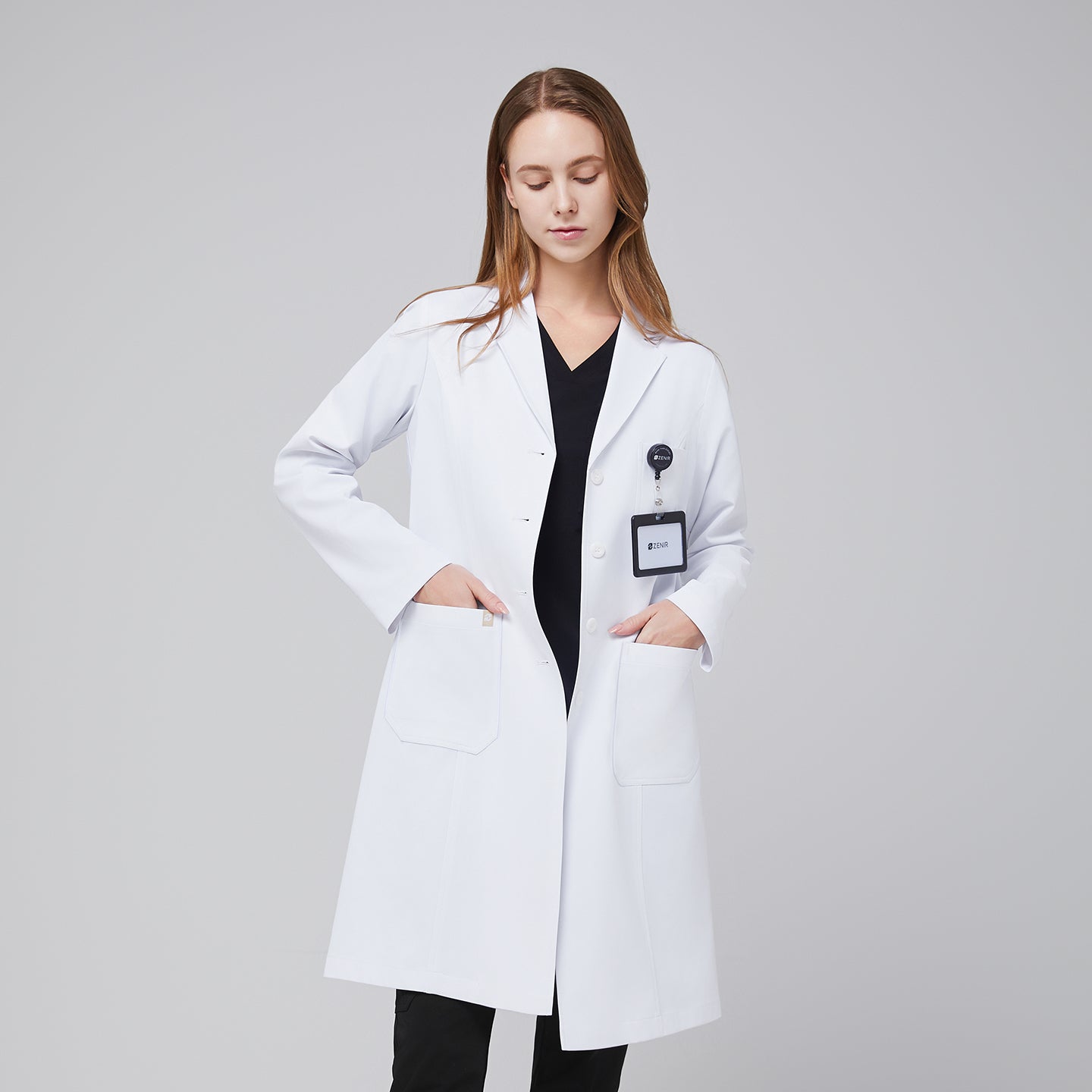 Image of Model facing forward looking down, with a white long lab coat and black scrub top and bottom under, with her hands in her lab coat pocket,White
