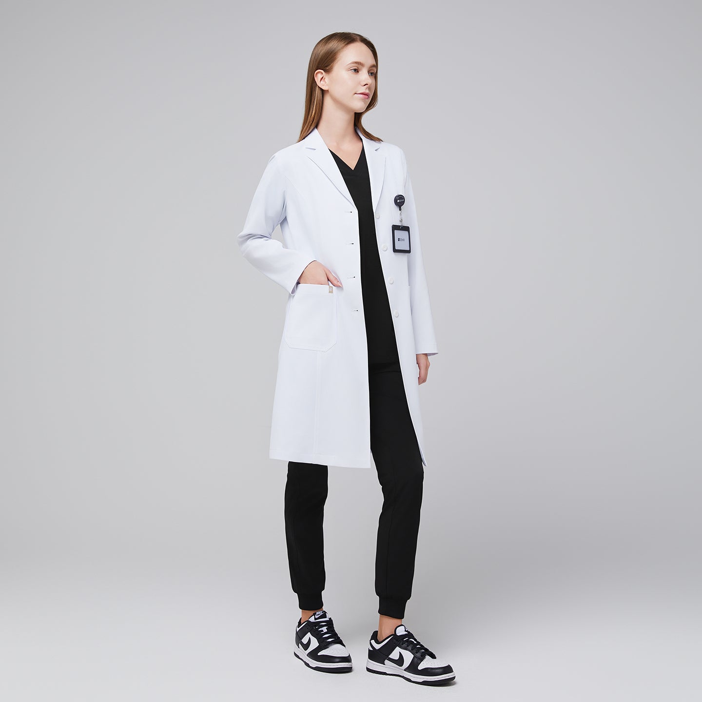 Model facing sideways, with a white long lab coat and black scrub top, black scrub pants, and black sneakers, with her hand in her lab coat pocket,White