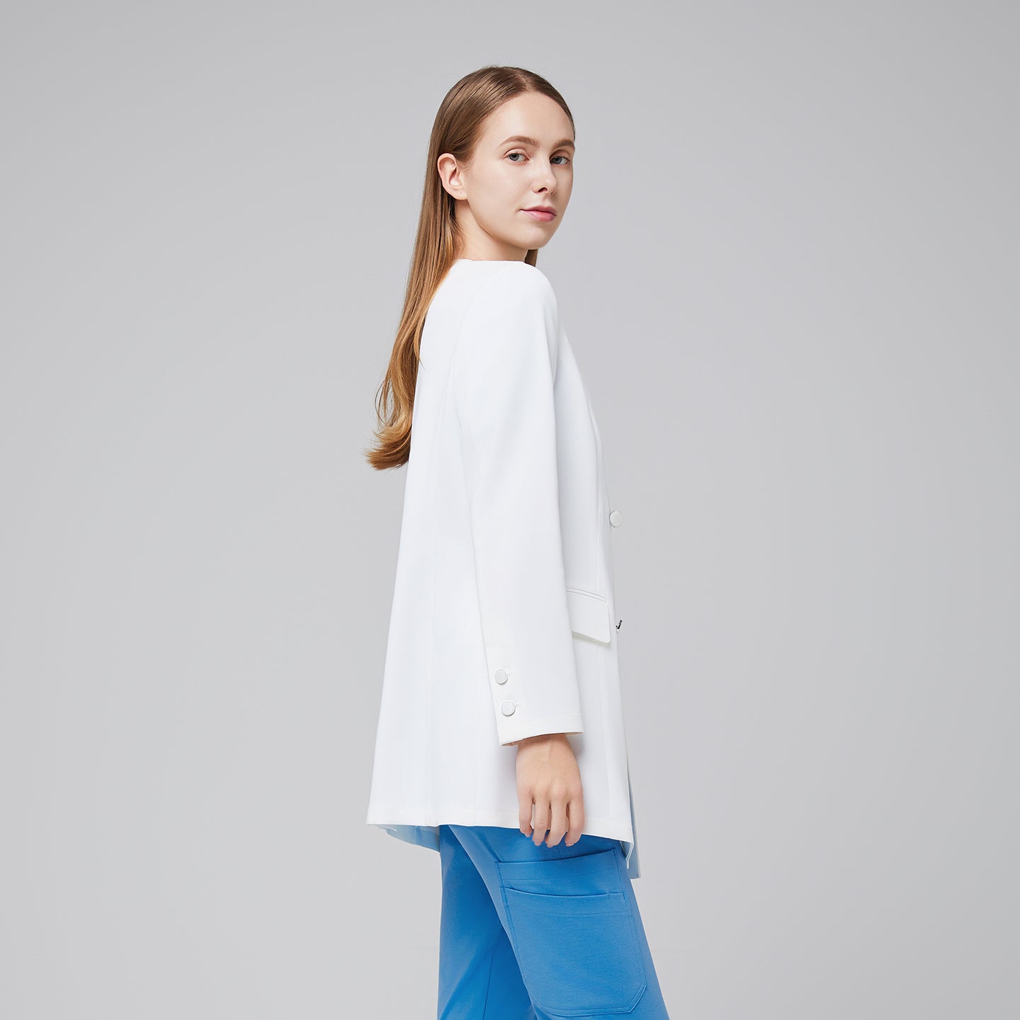 Profile shot of a woman wearing a white long lab coat with front pockets and four-button closure, paired with blue scrub pants,White