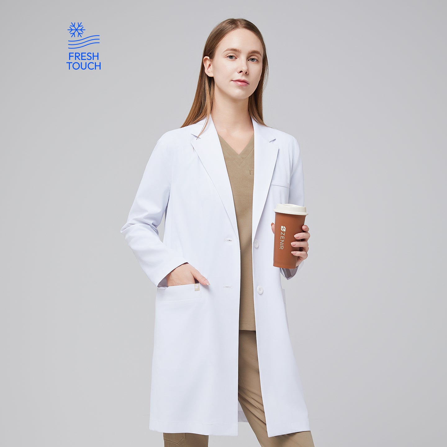 Image of Model facing forward with a hand in her pocket, wearing a white long lab coat with chest and side pockets, with a sand beige scrub top and bottom under, holding a brown tumbler,White