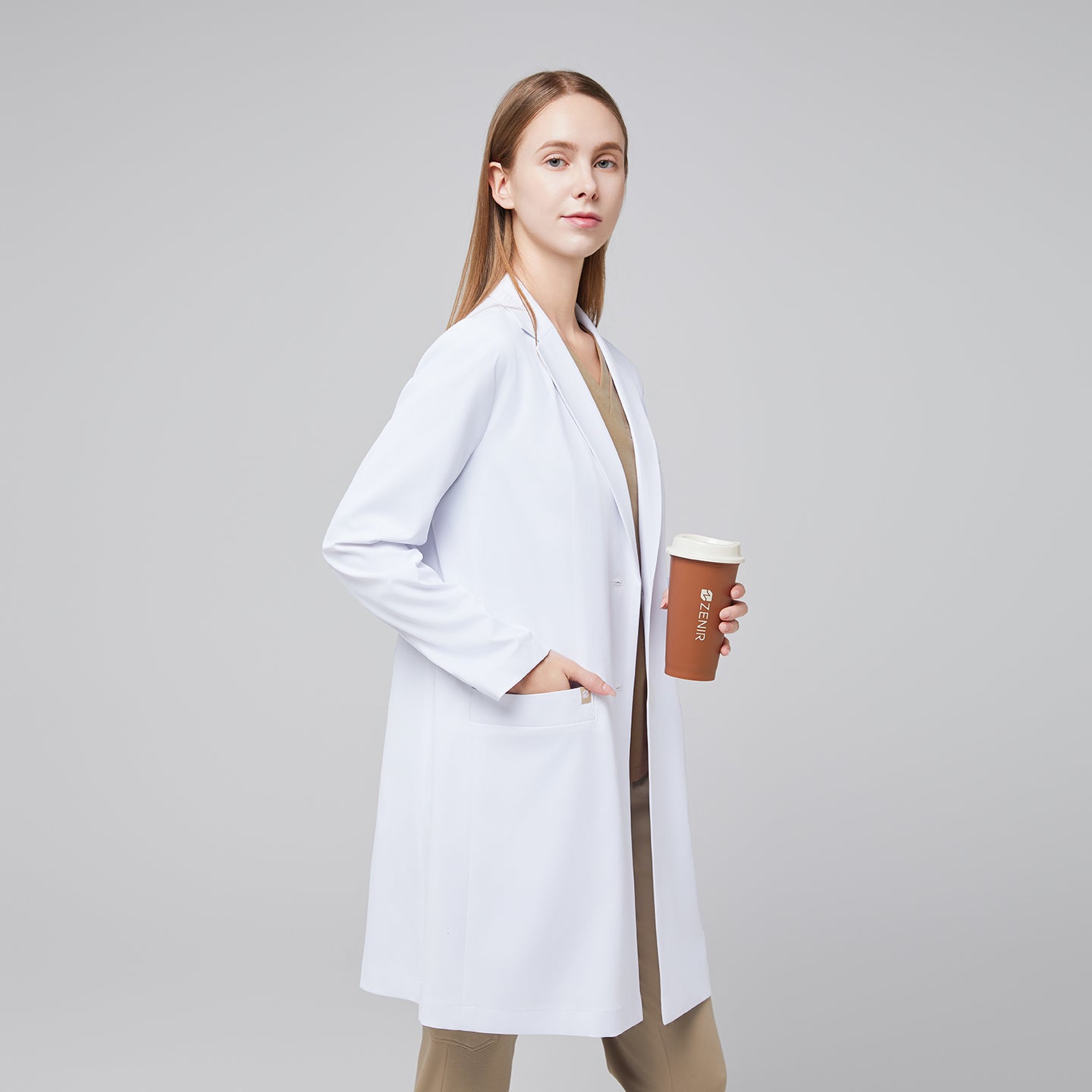 Image of Model standing to the side with a hand in her pocket, wearing a white long lab coat with chest and side pockets, with a sand beige scrub top and bottom under, holding a brown tumbler,White