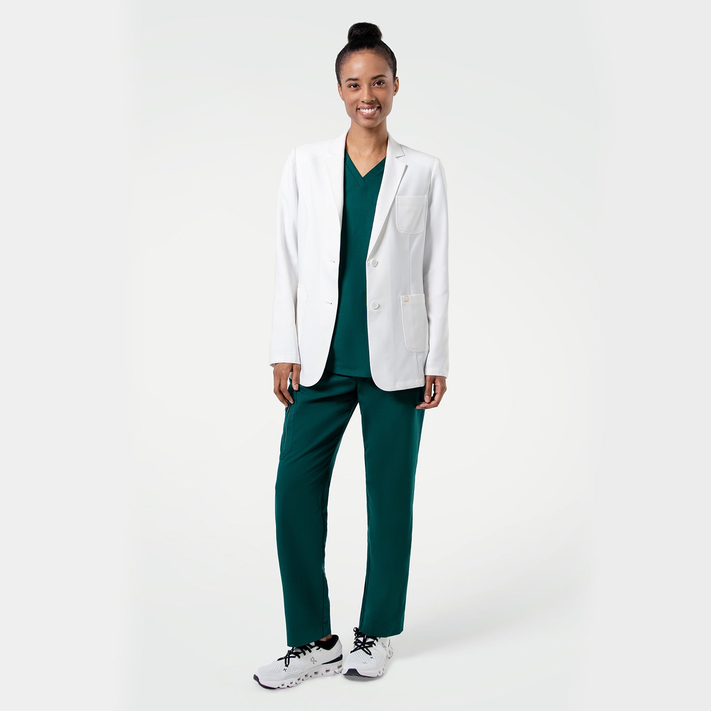 Image of Standing pose of the model wearing the Women's LCW-101 Lab Coat over pine green scrubs, smiling with one leg slightly bent