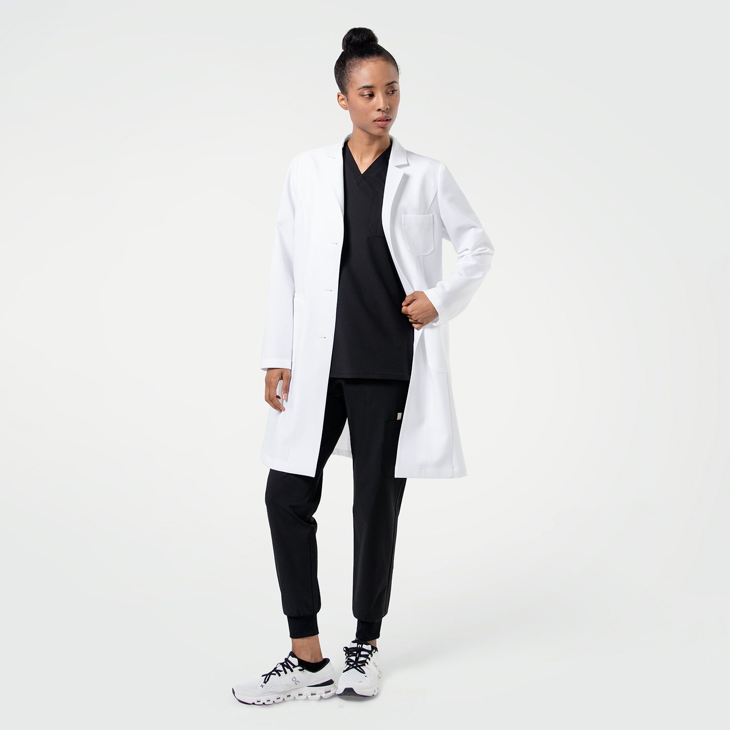 Image of Woman wearing the Women's LCW-102 Lab Coat in White over medical scrubs, standing facing forward while slightly looking to the side and holding one side of the coat
