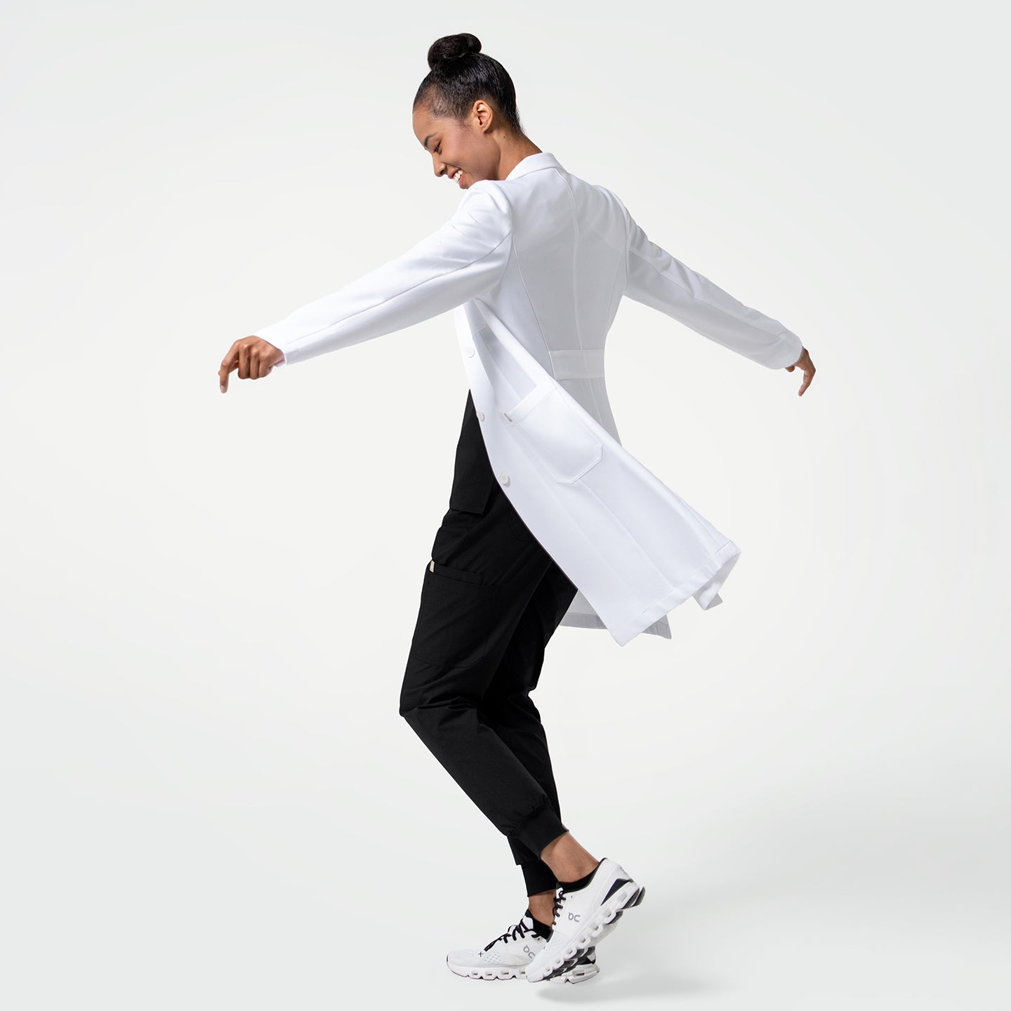 Image of Woman_wearing_the_Women_s_LCW-102_Lab_Coat_in_White_over_medical_scrubs_full_body_shot_captured_mid-spin_showing_a_dynamic_side_view