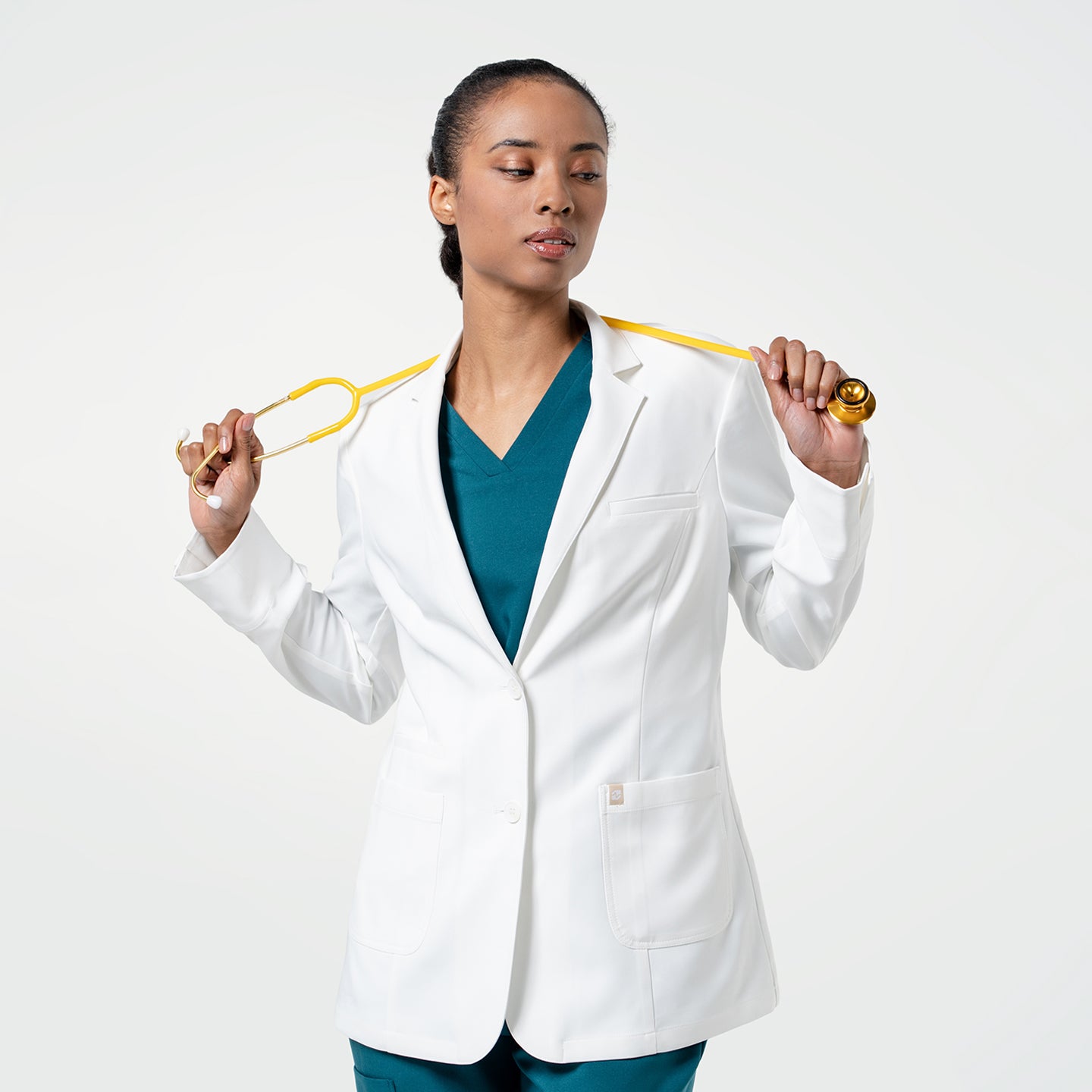 Image of With the coat buttoned, the model wearing the Women's LCW-04 Lab Coat in White over medical scrubs holds a yellow stethoscope around her neck with both hands, upper body view