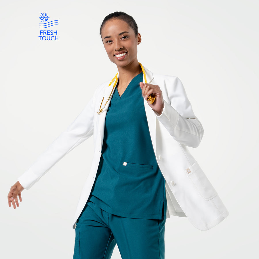 Smiling, the model in the Women's LCW-04 Lab Coat in White over medical scrubs moves with a yellow stethoscope around her neck, upper body view