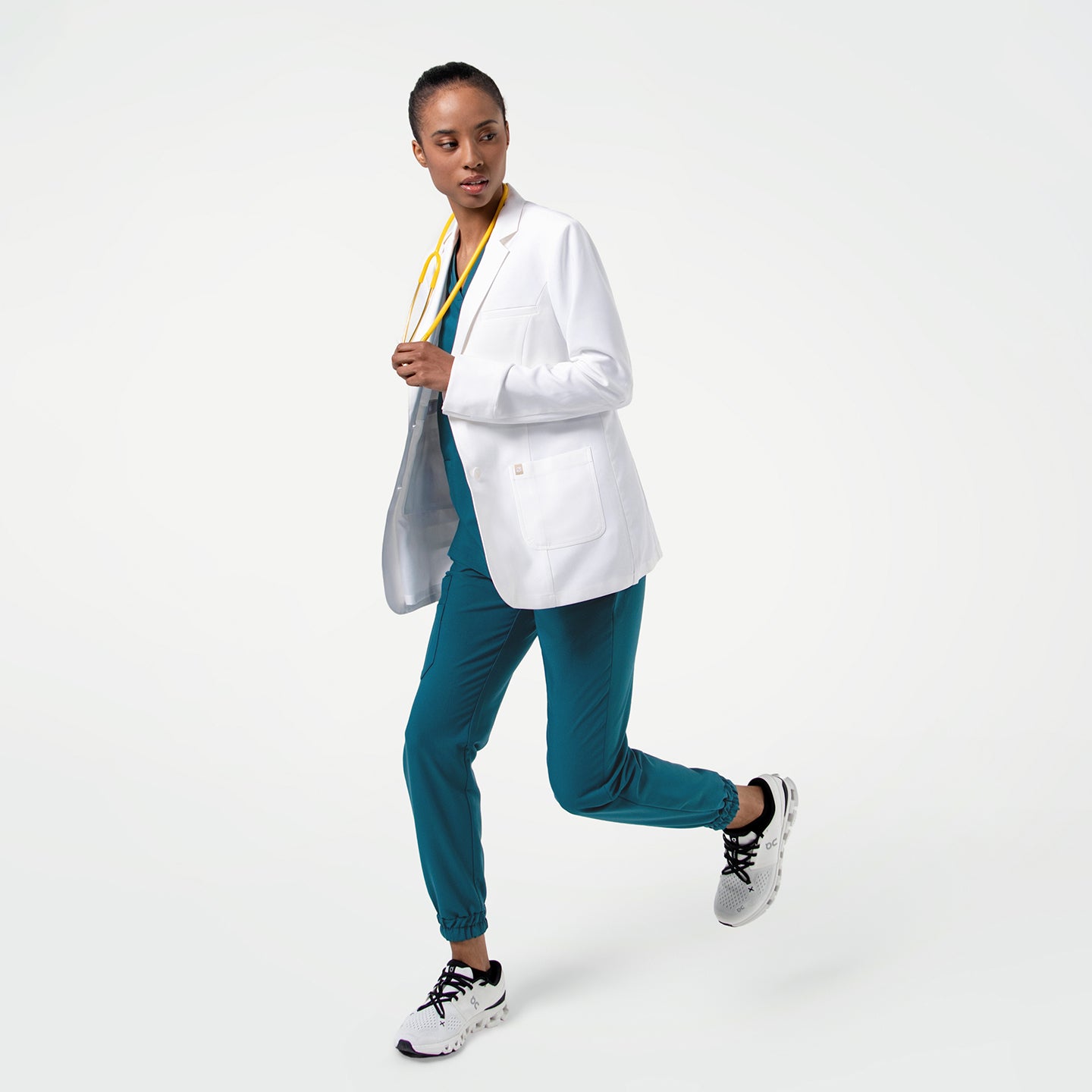 Image of Captured in motion, the model wears the Women's LCW-04 Lab Coat in White over medical scrubs, running toward the camera with her body slightly angled and a yellow stethoscope around her neck