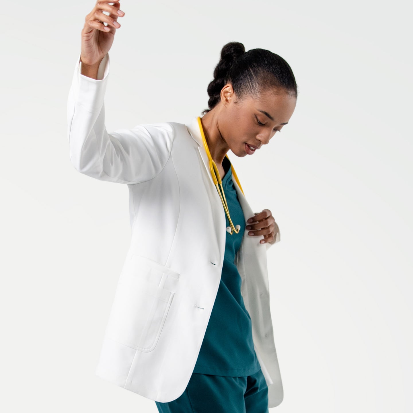 Image of In a dynamic pose, the model wears the Women's LCW-04 Lab Coat in White over medical scrubs, with a yellow stethoscope around her neck and one hand slightly pulling the coat aside