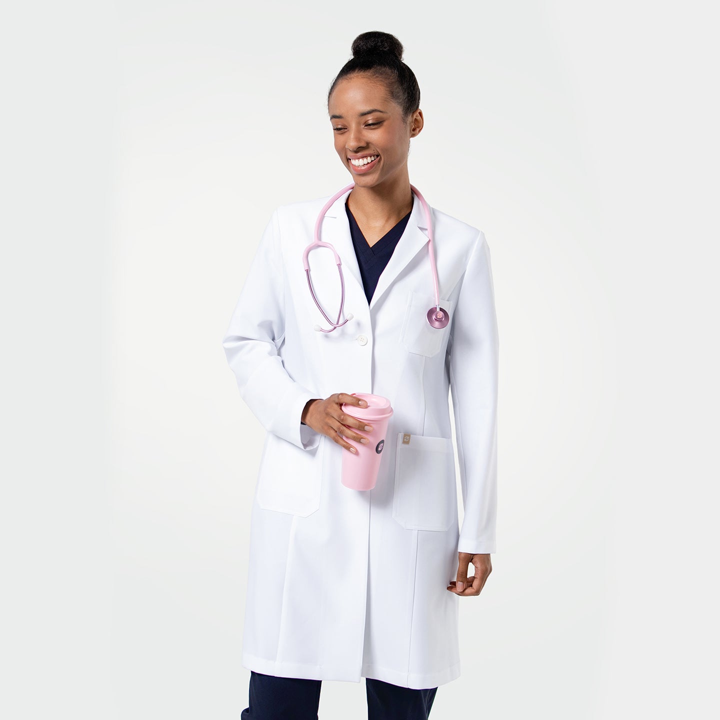 Image of Healthcare professional wearing a long white lab coat (LCW-203) over navy scrubs, holding a pink tumbler with a stethoscope around the neck, standing against a light background.