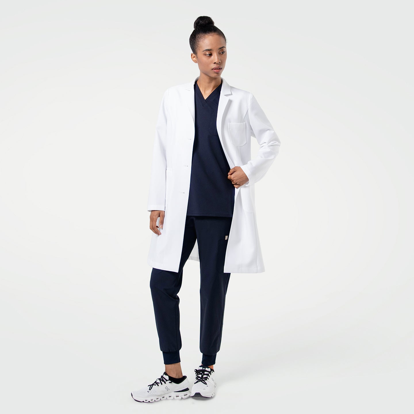 Image of LCW-203 long white lab coat shown on a model wearing navy scrubs, featuring a clean tailored fit and front patch pockets.