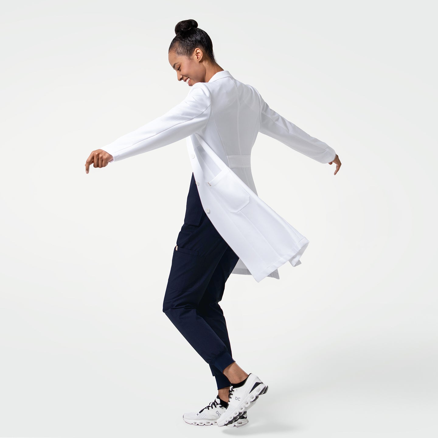 Image of A woman wearing a crisp white lab coat is shown mid-movement, arms gracefully extended as the coat flares behind her. The tailored back panel, clean seams, and structured silhouette highlight the coat’s professional, polished look.