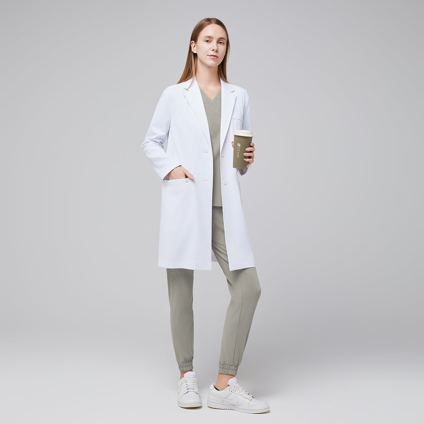 Image of A clean, mid-length white lab coat with a tailored silhouette, featuring a notched lapel, two spacious front pockets, and a streamlined fit worn over light sage scrubs for a polished professional look.