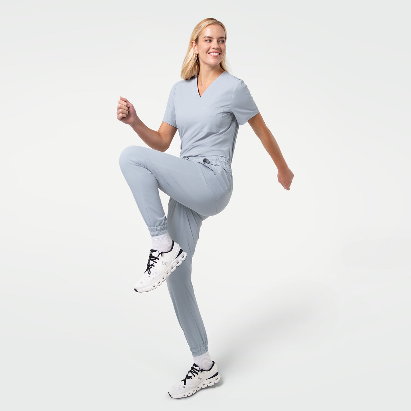 Image of Captured mid-motion with one knee lifted, the model wears a Cool Gray V-neck scrub top and matching jogger-style scrub pants, paired with white sneakers, smiling confidently against a clean light background.