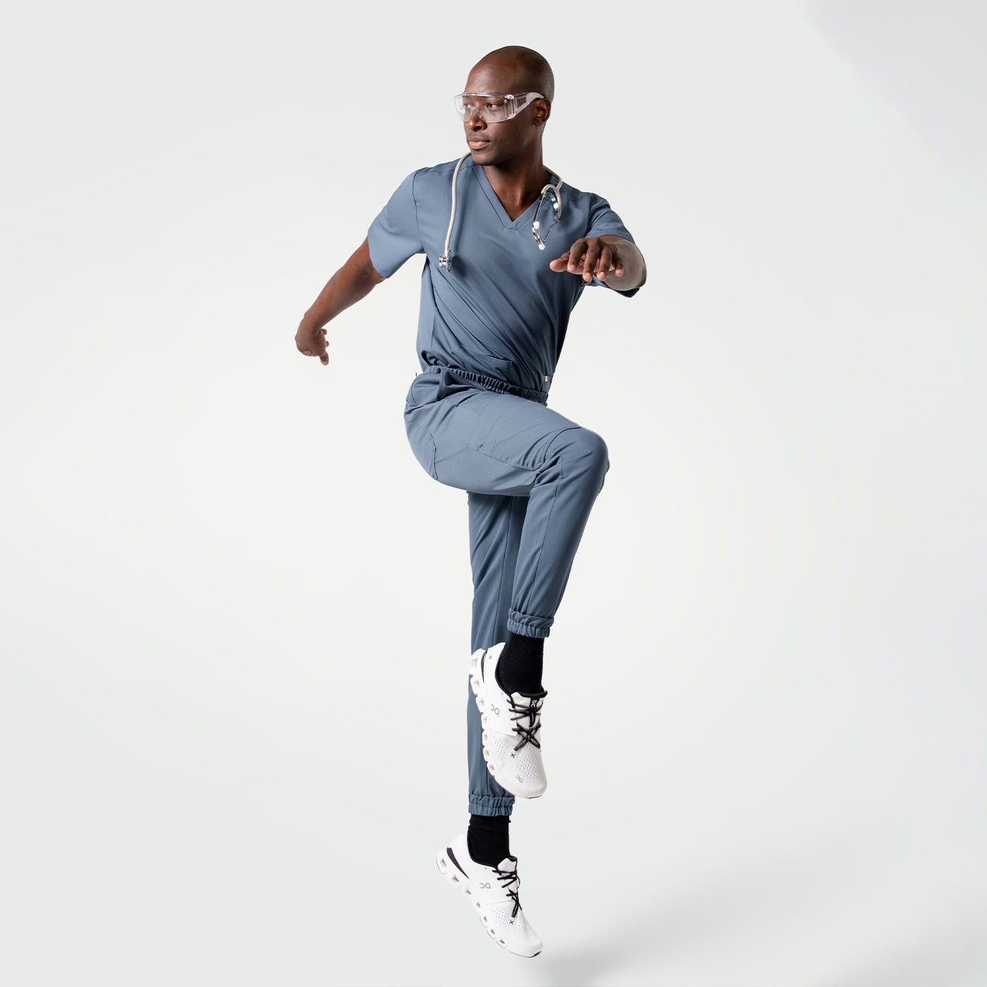 Image of Model performing a dynamic jump pose while wearing the Smoky Blue scrub set with a stethoscope around the neck.