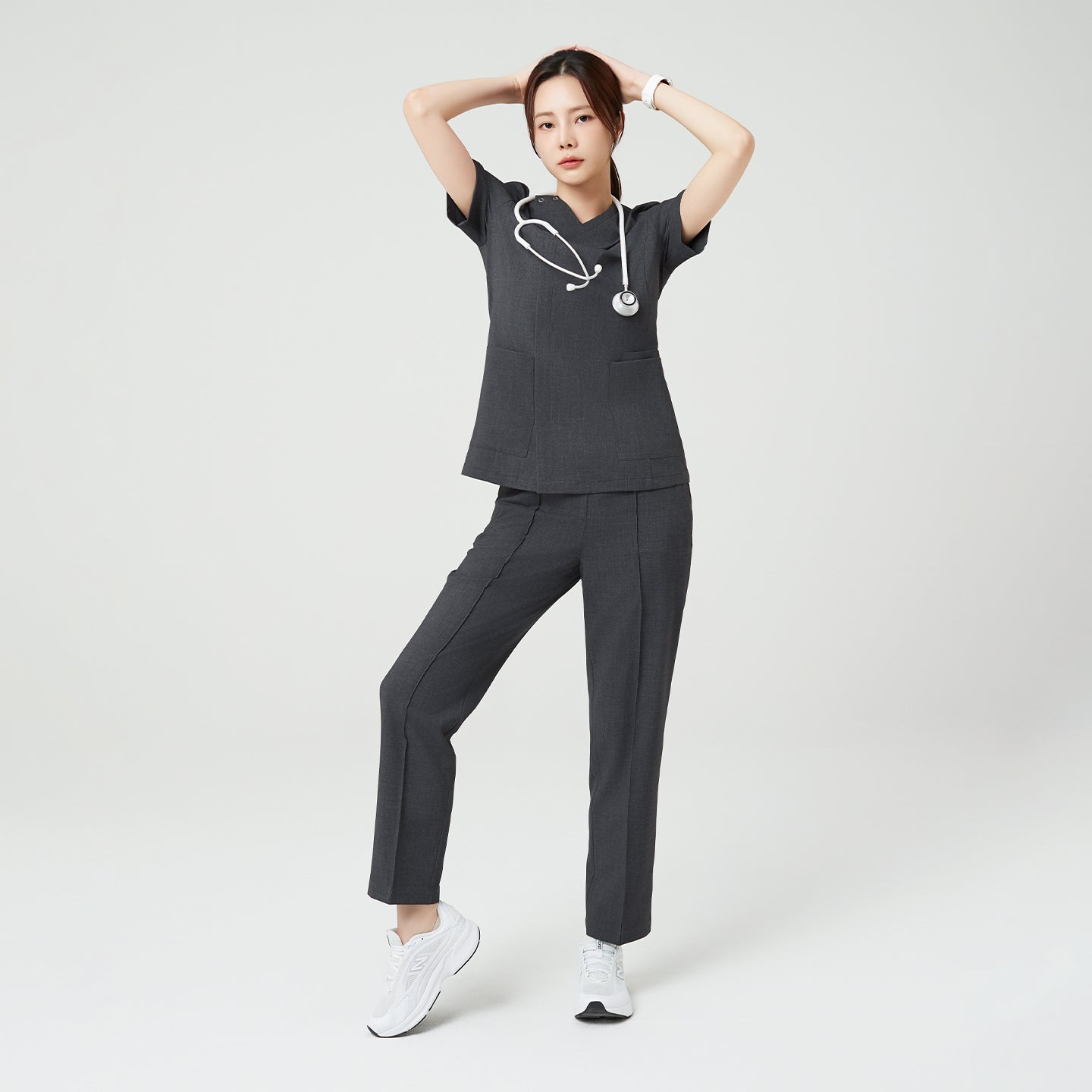 The Charcoal Gray OS-228 shows the model with arms raised slightly behind the head, highlighting the scrub’s flexibility, and relaxed fit.