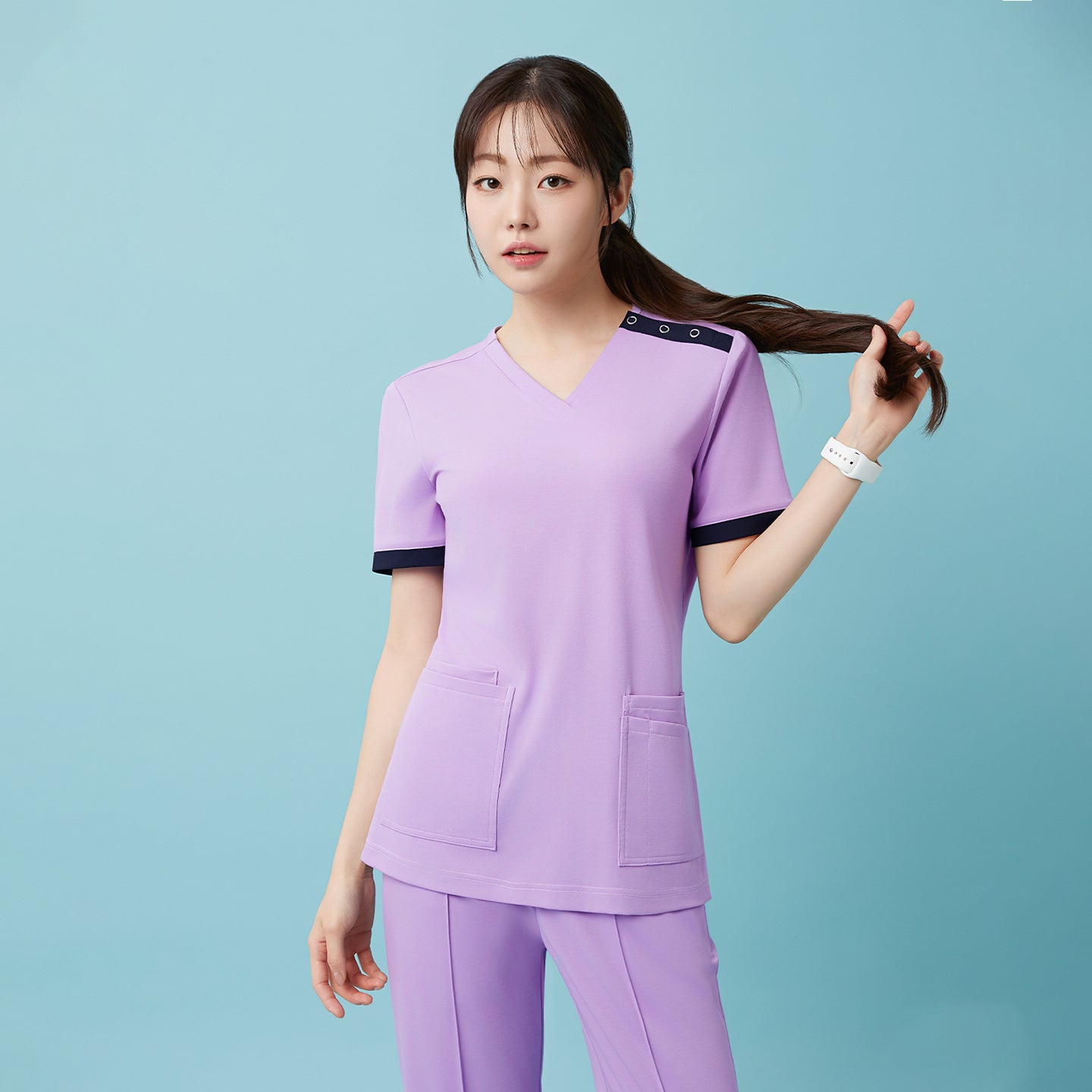 Image of Wearing the Lavender OS-230 scrub set, the model poses naturally while holding her ponytail, highlighting the soft pastel tone and relaxed fit that exude a calm, professional charm.
