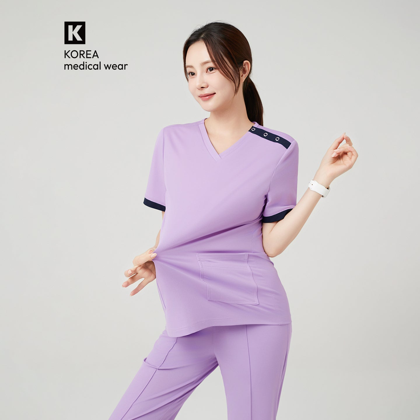 A female model is shown wearing The Uniform Lab OS-230 scrub top in Lavender, featuring a soft and stretchy fabric that she gently pulls forward to demonstrate its flexibility. The top includes contrasting navy snap-button details along the shoulder and matching navy trim at the sleeves, paired with coordinated lavender scrub pants for a clean, professional medical uniform look.