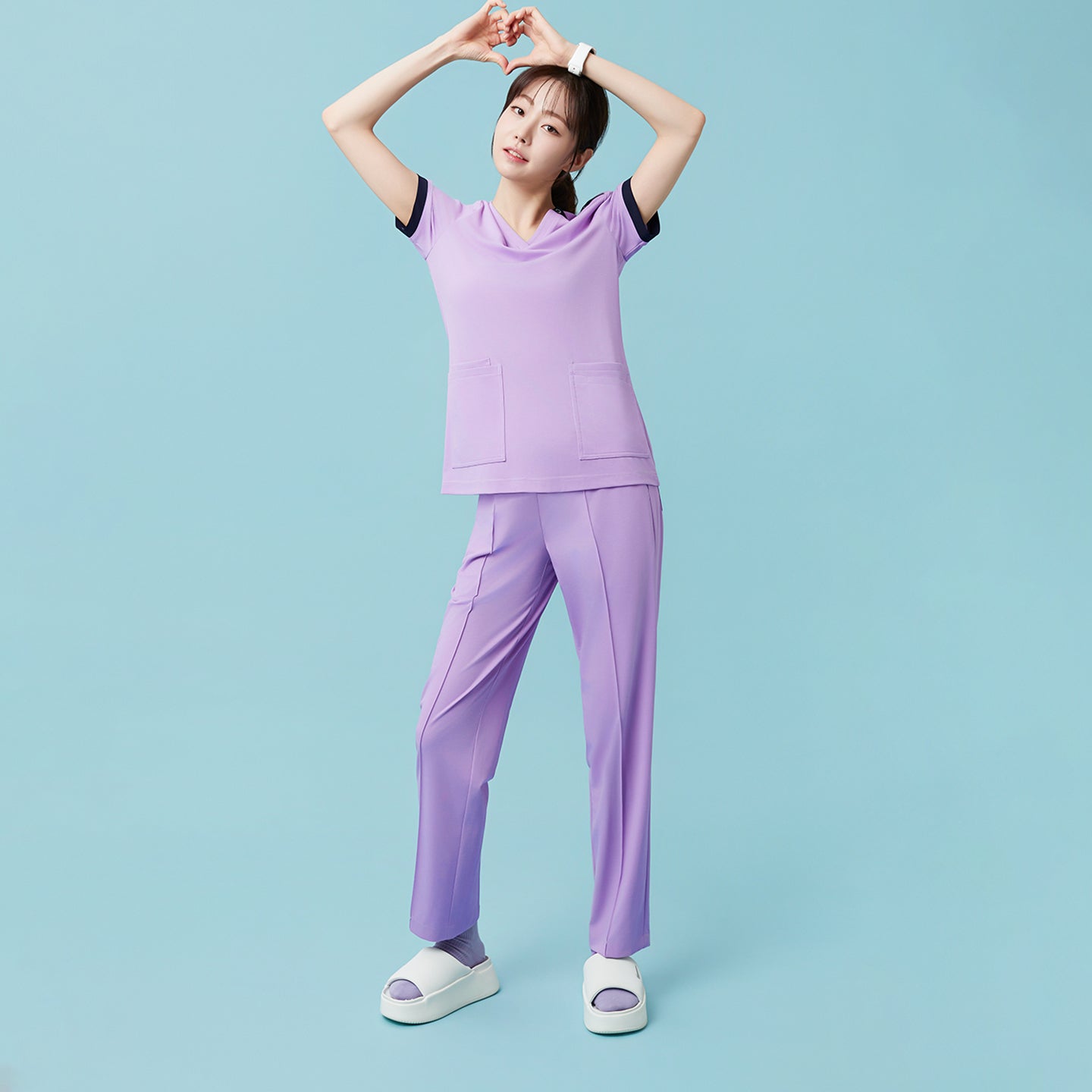 Image of With hands forming a heart above her head, the model adds a playful touch while showcasing the Lavender OS-230 scrub set’s flexible fit and lively pastel tone.