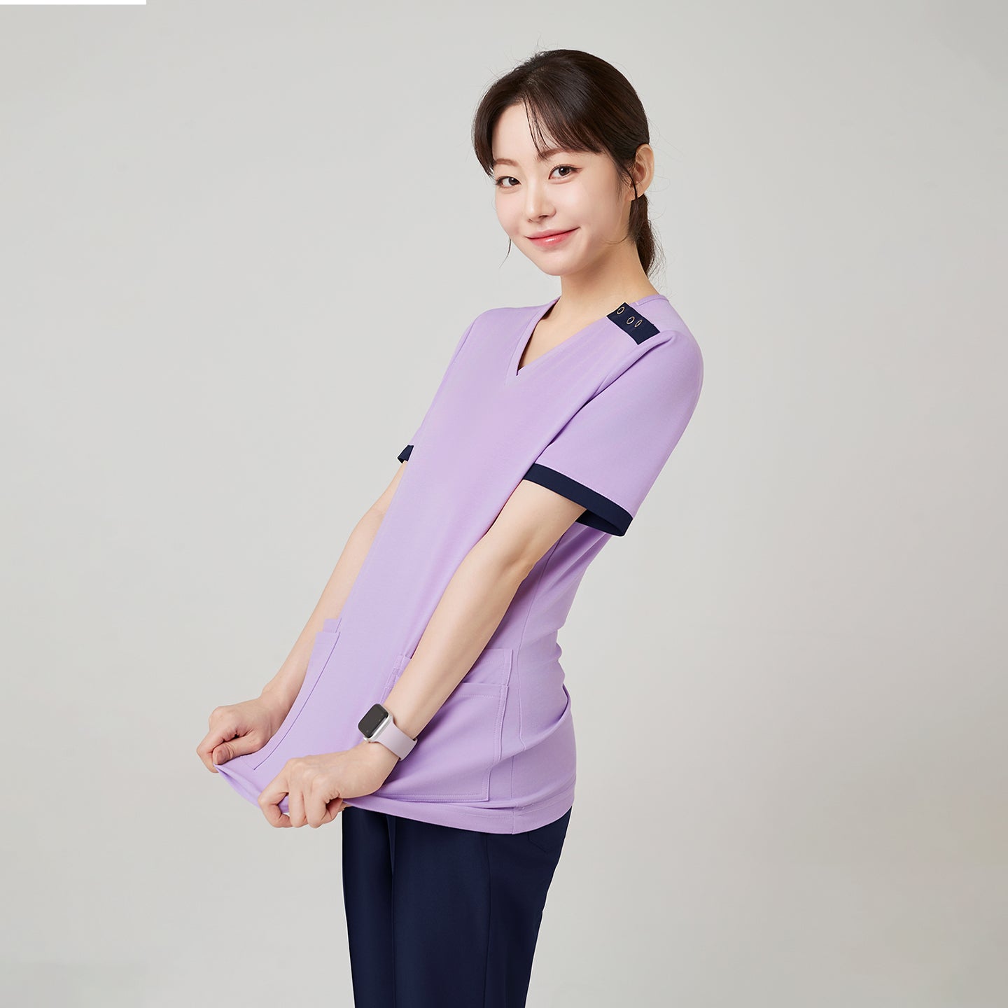 Image of The model gently stretches the fabric of the Lavender OS-230 scrub top, emphasizing its soft texture and comfortable stretch that moves easily with the body.