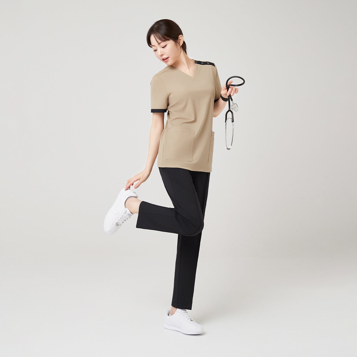 Image of Sand Beige OS-229 strikes a playful pose, lightly lifting one leg while holding a stethoscope, highlighting the scrub’s flexibility and modern two-tone design.