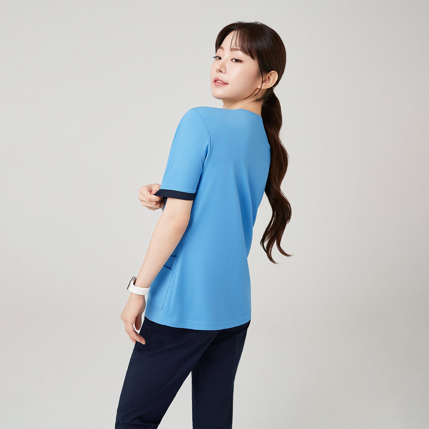 The model turns slightly to reveal the back view of the Sky Blue OS-231 scrub top, showing its clean lines and tailored fit that enhance both comfort and a polished silhouette.