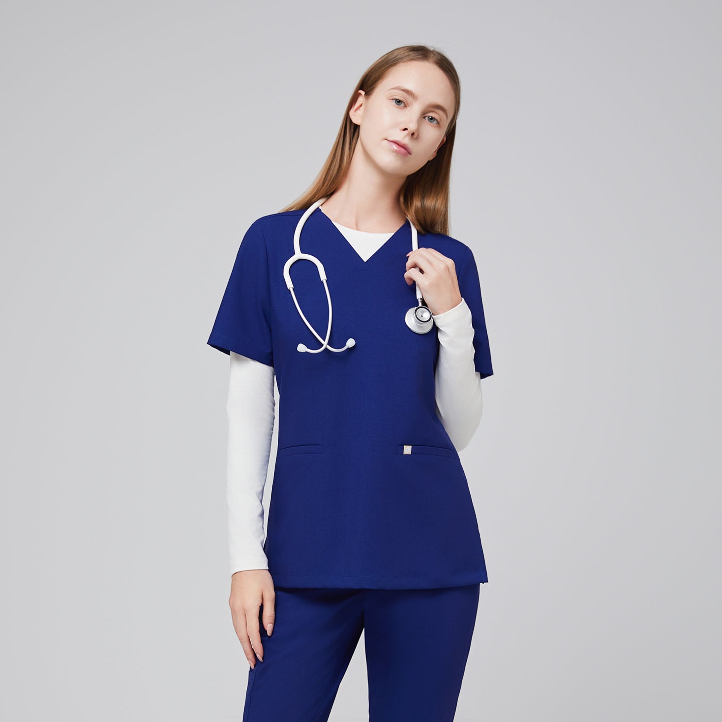 Image of Model wearing a navy blue double pocket V-neck scrub top layered over a white long sleeve shirt, with a stethoscope around the neck,Admiral Blue