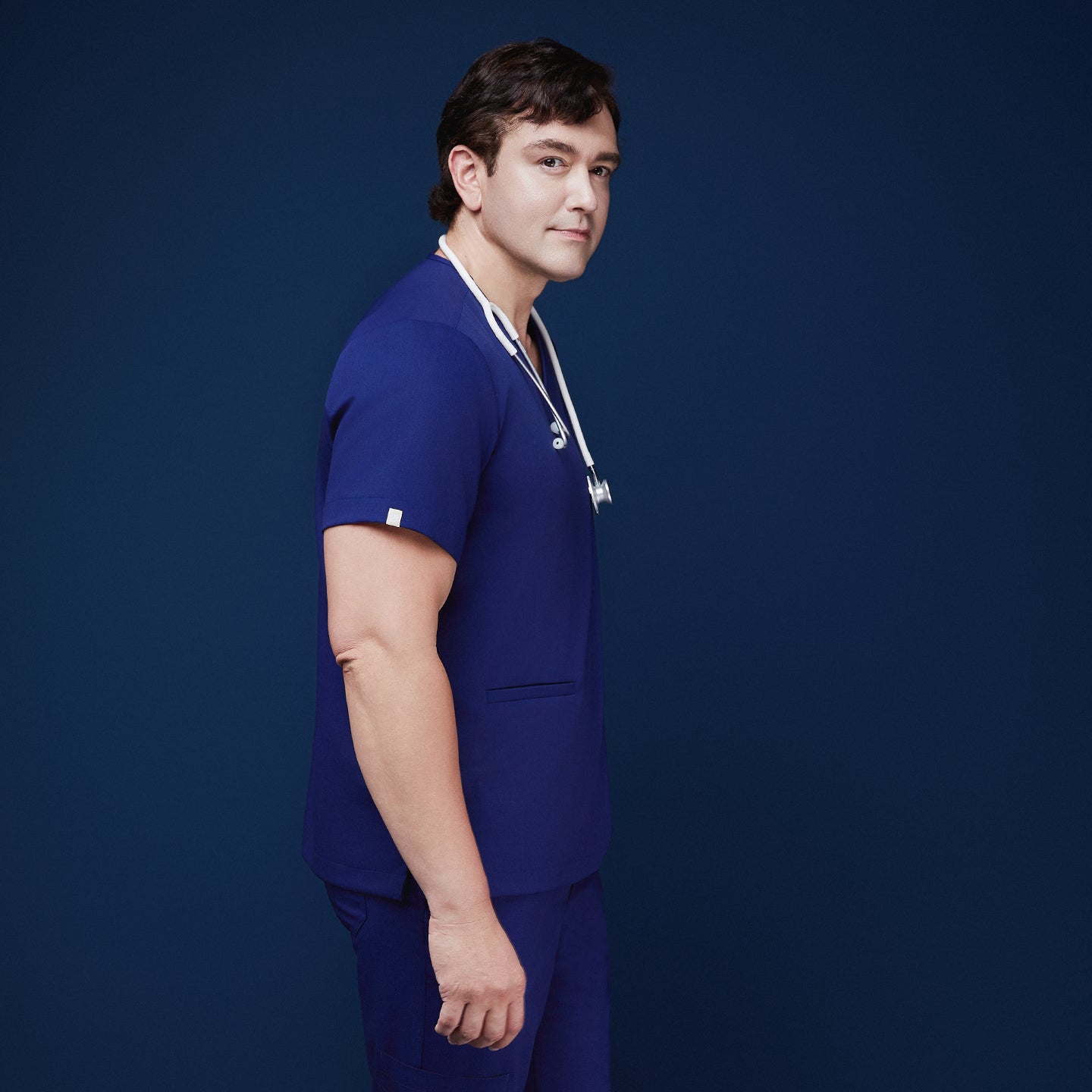 Image of Man wearing admiral blue scrubs with V-neck and double pockets, holding a white stethoscope around his neck,Admiral Blue
