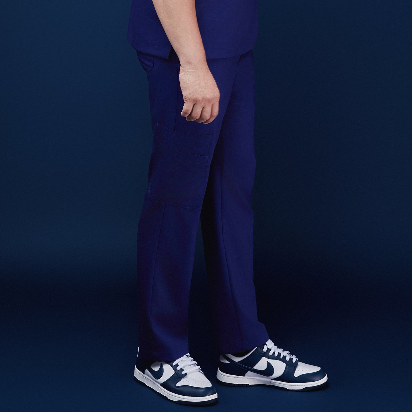 Image of Side view of model wearing admiral blue scrub pants with a pocket design, paired with navy and white sneakers, against a navy backdrop,Admiral Blue