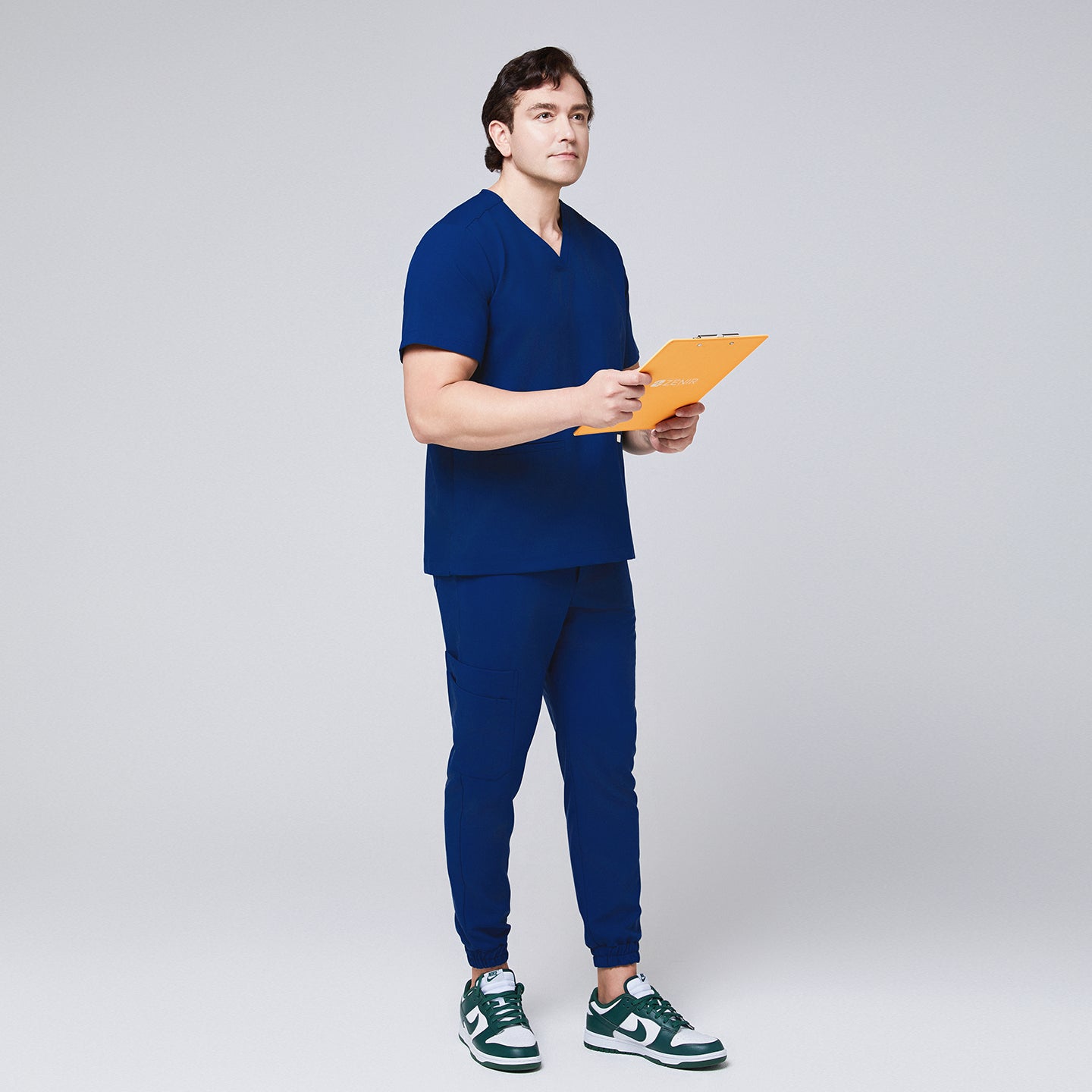Image of Full-body view of a man in Zenir Admiral Blue jogger scrub pants and matching top, holding a yellow clipboard, styled with green sneakers, Admiral Blue