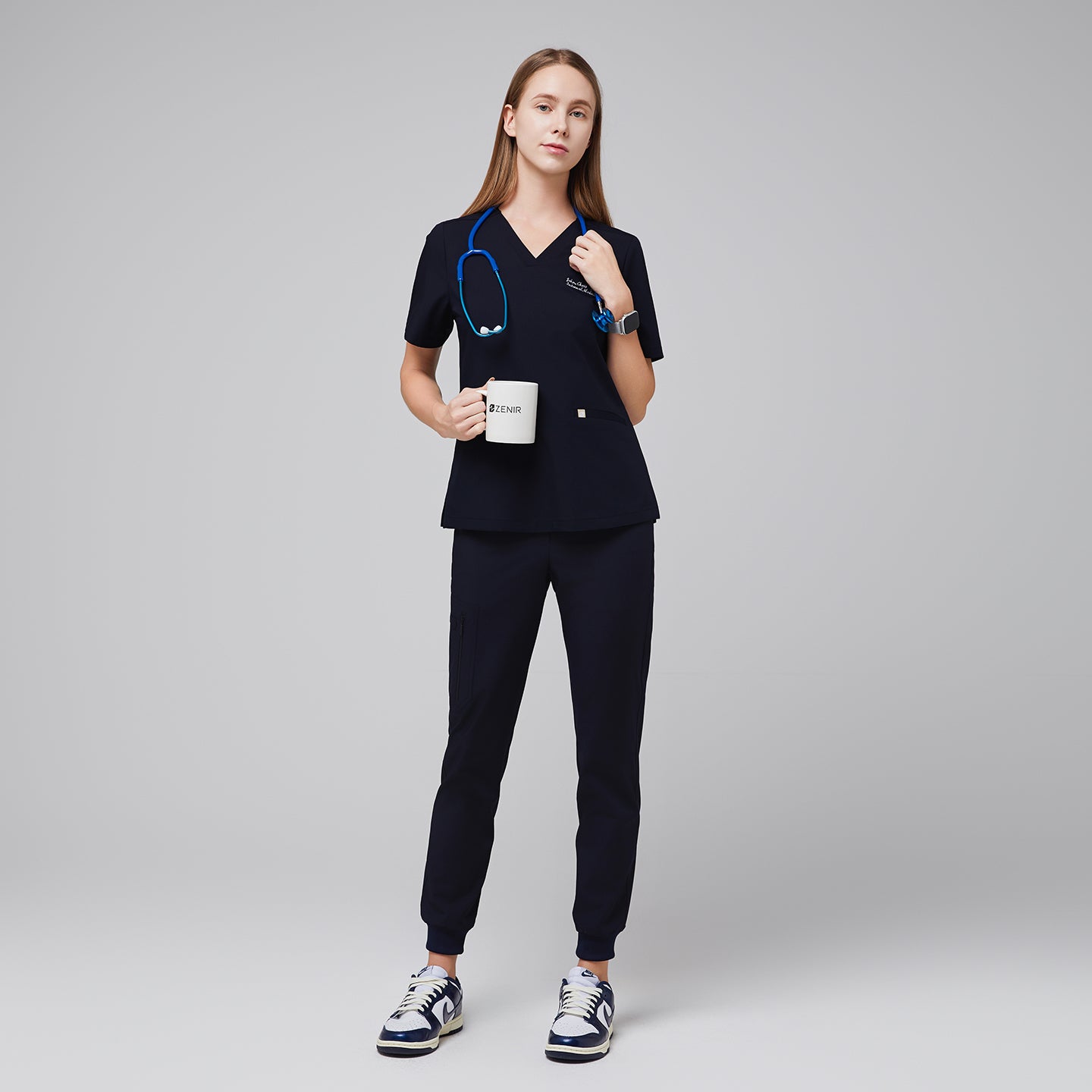 Image of Model in an eco navy double pocket scrub top with a blue stethoscope, holding a mug, and wearing matching jogger scrub pants and sneakers,Eco Navy